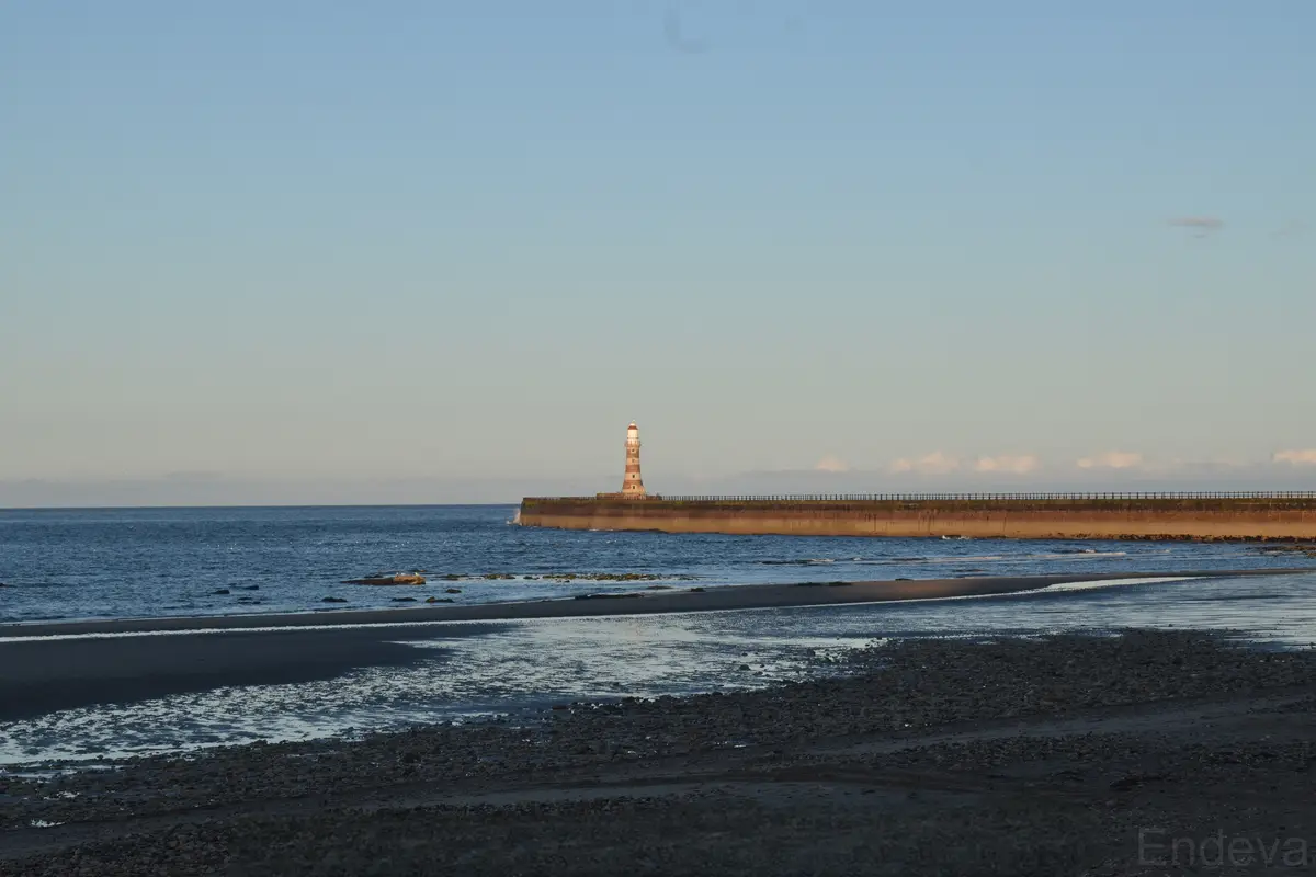 A scenic photo of a lighthouse standing at the end of a long pier extending into the ocean, with a wet, rocky beach in the…