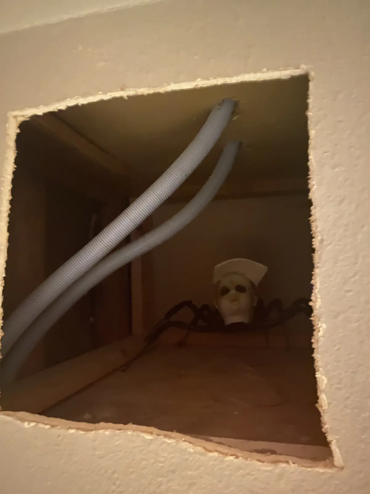 A square hole cut into a wall reveals a dark crawlspace where a nurse mask with black spider legs is positioned behind two…