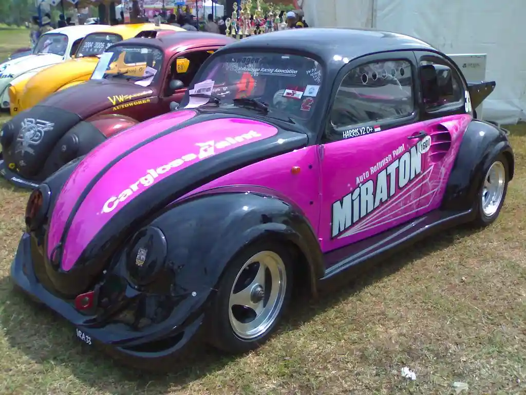 A modified black and pink Volkswagen Beetle race car with sponsor decals like 'Cargloss Paint' and 'MiRATON' is parked at an…