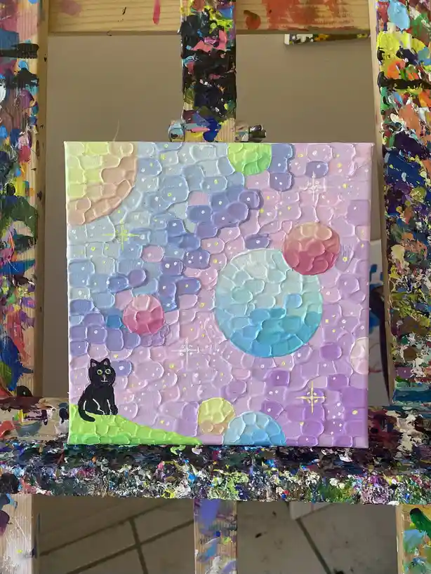 A small, textured painting on canvas depicts a black cat sitting on a green hill under a pastel-colored, starry sky with…