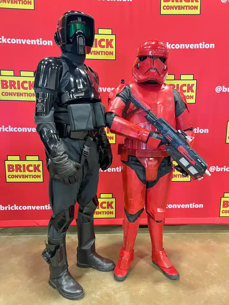 Two people in full Star Wars cosplay stand side-by-side at a Brick Convention backdrop. The person on the left wears a black…