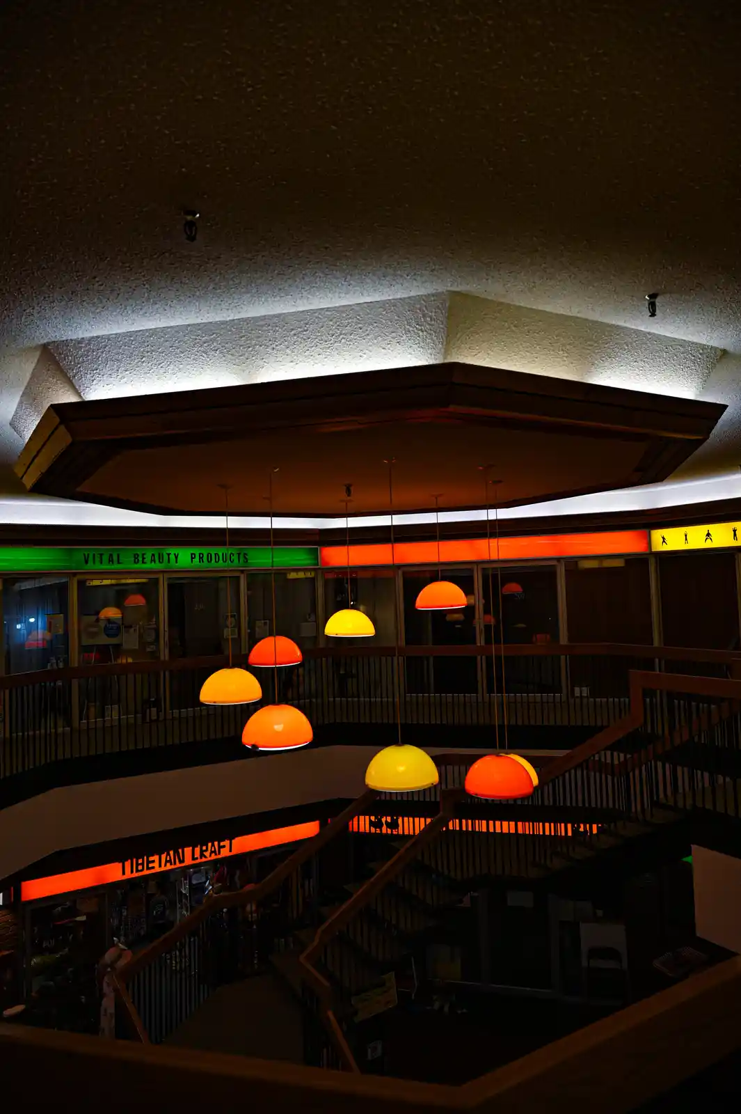 A dimly lit, multi-level indoor space with hanging orange and yellow dome lights, featuring storefronts labeled 'VITAL…